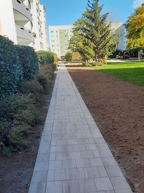 Das Bild zeigt einen Gehweg. Der Gehweg wurde neu gebaut. Der Gehweg gehört zu einem Innenhof. Um den Innenhof stehen Wohnhäuser.
