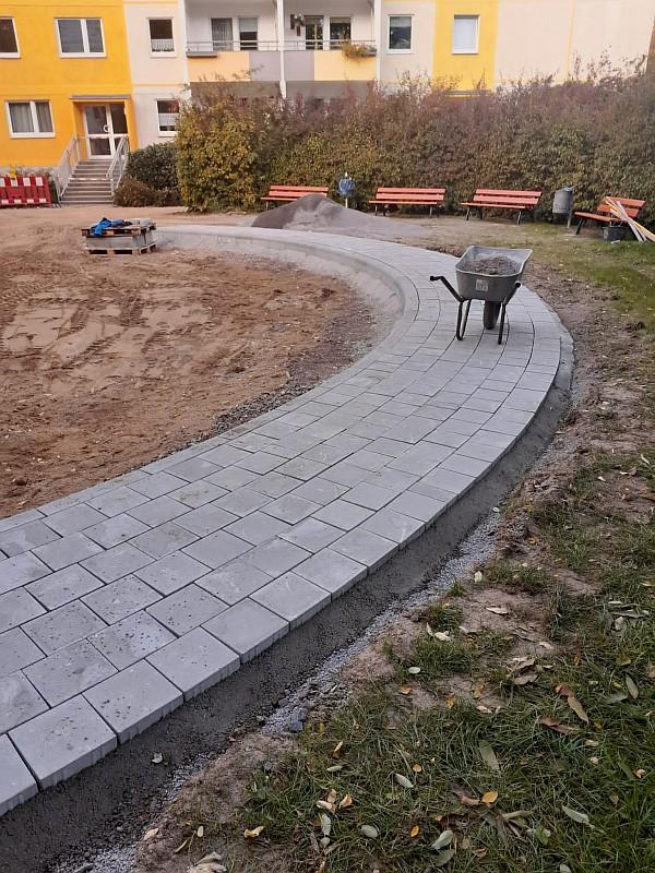 Das Bild zeigt einen Weg. Der Weg wurde neu gebaut. Der Weg gehört zu einem Sandkasten. Der Sandkasten befindet sich in einem Innenhof. Um den Innenhof stehen Wohnhäuser.