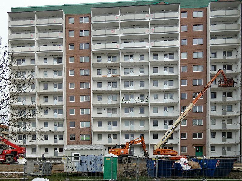 Auf dem Bild sieht man einen Kran. Der hat Menschen auf einen Balkon befördert. Die bauen die Absturzsicherung ab. Die Balkone sollen abgerissen werden.