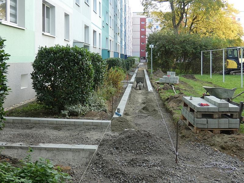 Das Bild zeigt den Bau eines Gehweges. Der Gehweg wird in einem Innenhof errichtet. Um den Innenhof stehen Wohnhäuser.